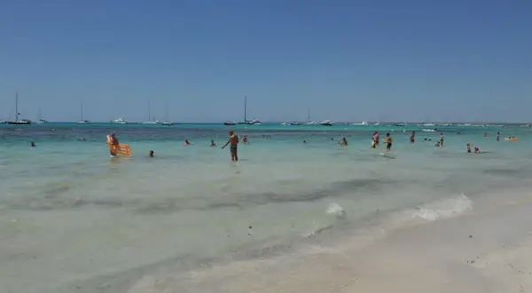 Słoneczny dzień na plaży Es Trenc. Ludzie kąpią się w turkusowej wodzie, na horyzoncie widać żaglówki.