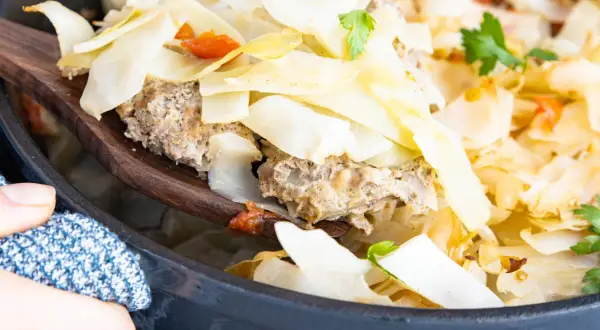 Ein Holzlöffel hebt eine Portion Krauttopf mit Hackfleisch aus einem Topf. Das Gericht ist im Ofen gebacken und mit Petersilie garniert.