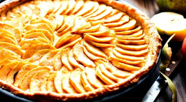 Ein wunderschönes Apfeltarte Rezept, das mit dünn geschnittenen Äpfeln belegt ist. Daneben liegen frische Äpfel und ein Messer.