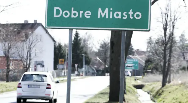 Zielony znak miejscowości "Dobre Miasto" wita podróżnych. W tle biały samochód i domy.