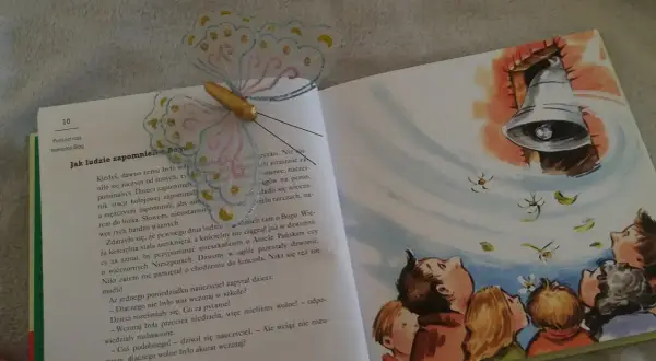 Otwarta książka z ilustracją dzwonu i dzieci patrzących w górę. Motylowa zakładka ozdabia stronę z tekstem o tym, jak ludzie zapomnieli o Bogu.