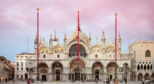 La maestosa Basilica di San Marco a Venezia, con le sue cupole dorate e mosaici scintillanti, si staglia contro un cielo rosa al tramonto.