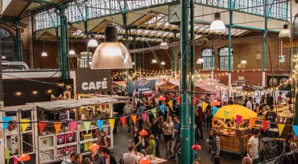 Buntes Treiben auf einem Street Food Festival in Berlin. Menschen schlendern zwischen Ständen mit Leckereien unter einer beeindruckenden Glasdachkonstruktion.