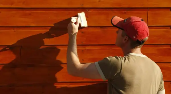 Un homme en casquette rouge est en train de peindre du bois brut extérieur avec un pinceau. La lumière du soleil projette une ombre nette sur le mur.