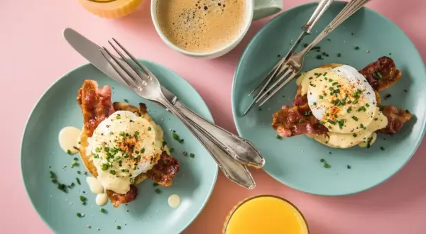 Zwei Teller mit **Egg Benedict**, garniert mit Speck und Schnittlauch, daneben Kaffee und Orangensaft.