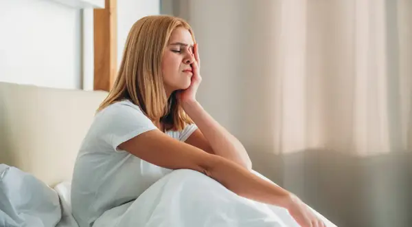 A young woman woke up with a headache, grimacing and holding her forehead as she sits in bed.