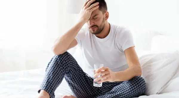 A man in pajamas sits on a bed, holding a glass of water and clutching his head, clearly sleeping off a hangover.