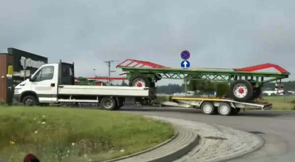 Transport przyczepy kempingowej na lawecie. Biała ciężarówka z platformą przewozi zielono-czerwoną przyczepę.