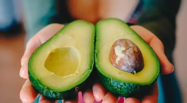 Manos sostienen un aguacate cortado por la mitad, listo para comer aguacate todos los días.