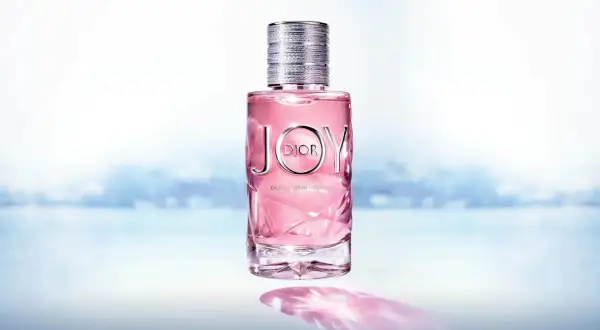 Różowy flakon Dior Joy 90ml z metalową nakrętką, na tle rozmytego, błękitnego krajobrazu.