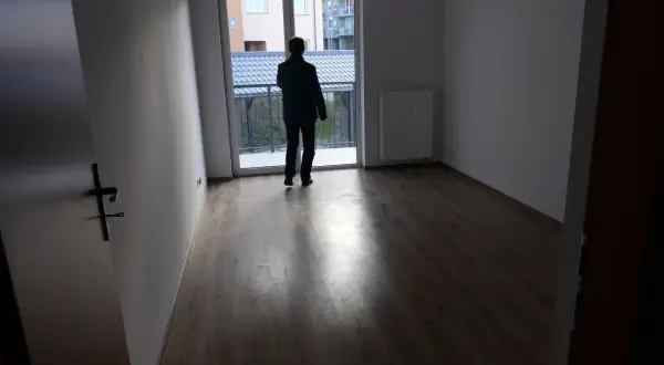 Mężczyzna w pustym mieszkaniu, zastanawia się, gdzie złożyć wniosek o mieszkanie komunalne. Widok na balkon i sąsiednie budynki.