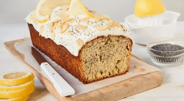Ein saftiger Zitronen Mohn Kuchen mit weißem Zuckerguss, Mandelsplittern und Zitronenscheiben. Daneben liegen frische Zitronenscheiben und Mohn.