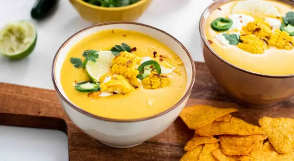 Zwei Schüsseln mit cremiger Mais-Suppe, garniert mit Maiskolben, Limette und Jalapeños, daneben Tortilla-Chips.