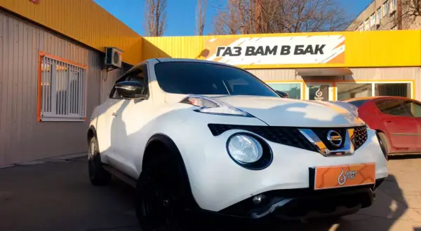 Biały Nissan Juke 1.6 benzyna z czarnymi felgami przed budynkiem z żółtym banerem "GAZ WAM W BAK".