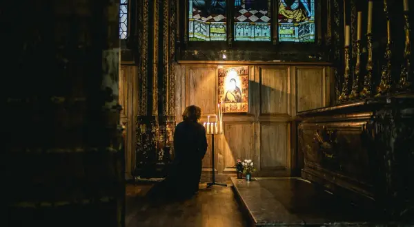 Osoba klęczy przed ikoną w ciemnym kościele, zapalając świece.
