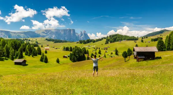 Ein Wanderer genießt die Aussicht auf eine Alm mit Berghütten und Wäldern. Perfekt für eine Mehrtageswanderung in den Dolomiten.