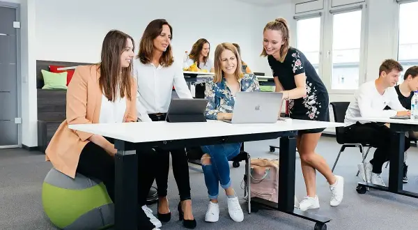 Vier junge Leute arbeiten an Laptops und Tablets. Sie diskutieren und lachen. Das Management Studium Dauer ist hier ein wichtiges Thema.