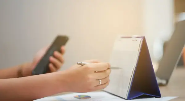 Ręka z długopisem zaznacza datę w kalendarzu, sprawdzając, czy 1 listopada jest dniem wolnym od pracy. Obok leży telefon.