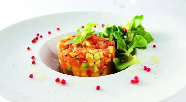 Délicieux tartare de truite aux agrumes, avocat et poivrons, garni de jeunes pousses et de baies roses sur une assiette blanche.