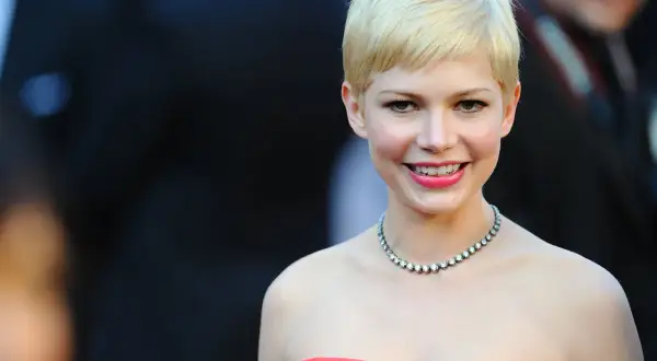 Eine Frau mit einem frechen Pixie-Cut und welligem Haar lächelt charmant. Sie trägt ein auffälliges Collier und ein rotes Kleid.