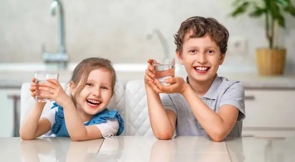 Uśmiechnięta dziewczynka i chłopiec trzymają szklanki z wodą. Cieszą się orzeźwiającym smakiem, jakby pili miękką wodę prosto ze źródła.