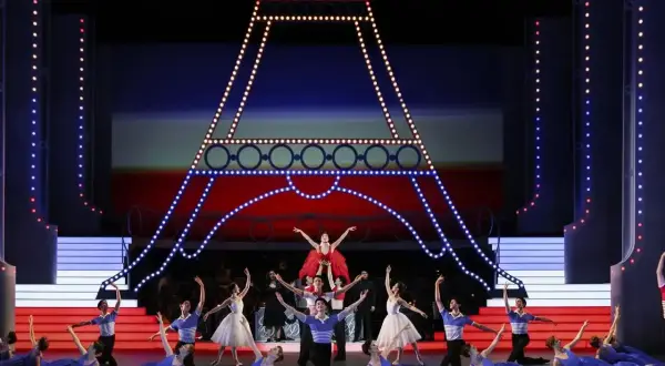 Tancerze baletowi na scenie z miniaturą Wieży Eiffla. W tle czerwono-niebieska scenografia, przypominająca flagę Francji. W powietrzu unosi się aria.