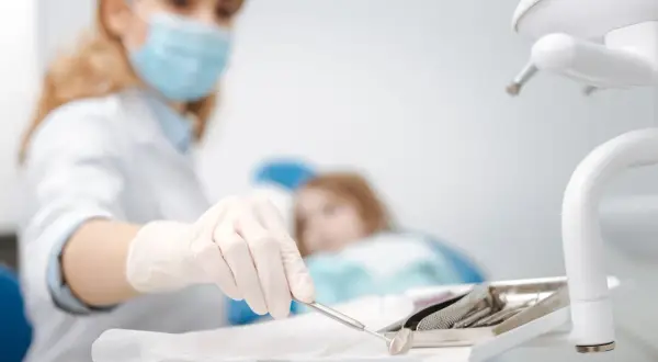 Dentystka w masce przygotowuje narzędzia do ekstrakcji zęba. W tle pacjentka na fotelu.