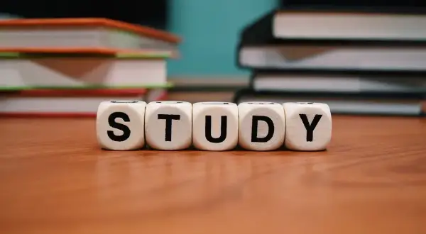 Białe kostki z literami "STUDY" na drewnianym blacie, w tle stosy książek.