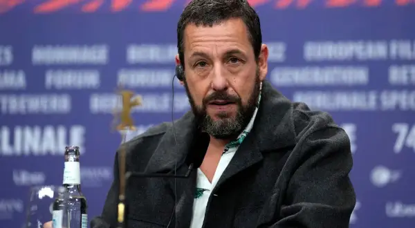 Adam Sandler, känd från många roliga filmer, sitter vid ett bord med en mikrofon och en vattenflaska.