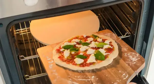 Przygotowania do dnia pizzy 2026: świeża pizza z mozzarellą i bazylią wsuwana na rozgrzany kamień w piekarniku.