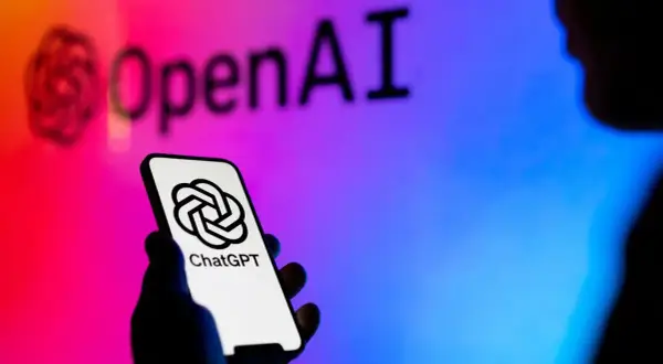 Dłoń trzyma smartfon z logo ChatGPT. W tle logo OpenAI, symbolizujące rozwój gpt sztuczna inteligencja.