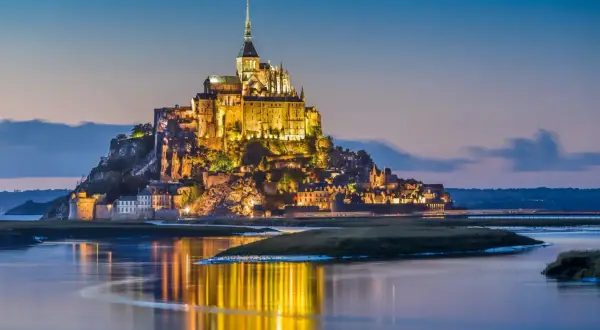 Mont Saint-Michel lyser upp i skymningen, en dröm för en semester i Frankrike. Vattnet speglar dess magiska ljus.