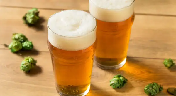 Dwa szklanki orzeźwiającego piwa hazy IPA z obfitą pianką, otoczone szyszkami chmielu na drewnianym blacie.