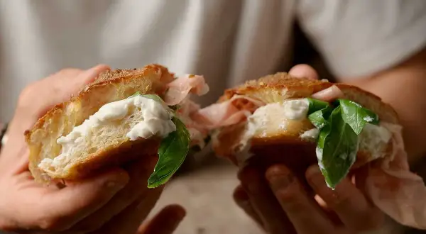 En person håller en delad focaccia med krämig ost, skinka och färsk basilika. En härlig focaccia pesto-macka!