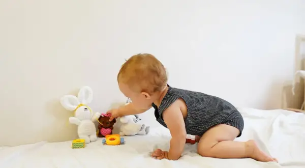 A baby explores Montessori baby activities, reaching for a teddy bear among soft toys on a white blanket.