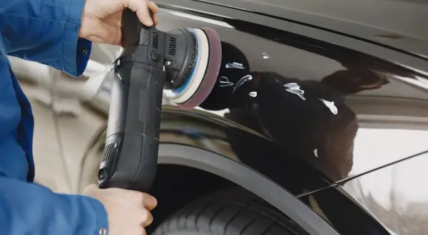 Osoba w niebieskiej kurtce poleruje czarne auto maszyną, pokazując jak usunąć rysy z auta.
