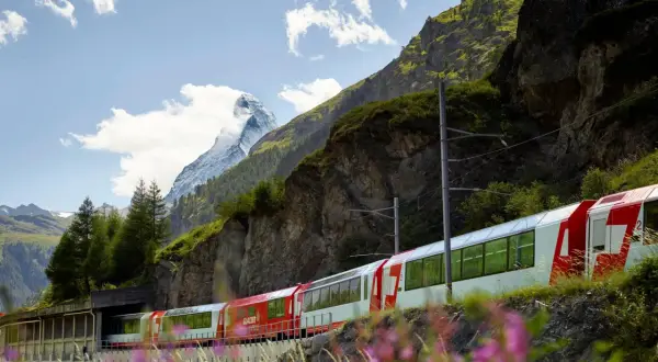 Czerwono-biały pociąg Glacier Express mija skaliste zbocze górskie z ośnieżonym szczytem w tle.