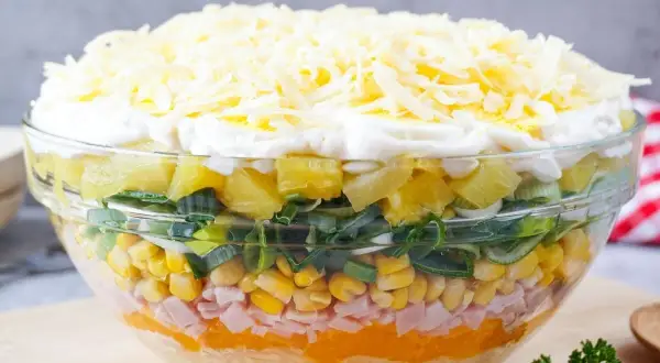 Schichtsalat mit Eisbergsalat, Mais, Schinken, Ananas, Lauch und Käse in einer Glasschüssel.