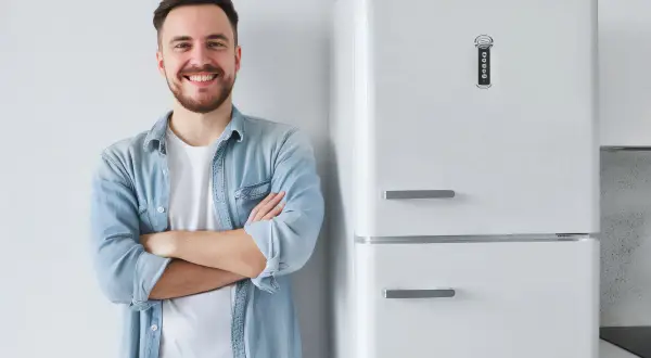 Uśmiechnięty mężczyzna w jeansowej koszuli stoi obok białej lodówki.