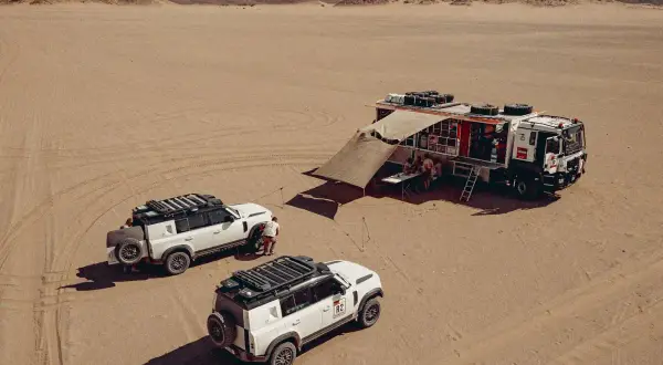 Un camion Dakar et deux 4x4 blancs dans le désert. L'équipe se réunit sous un auvent, prête pour l'aventure.