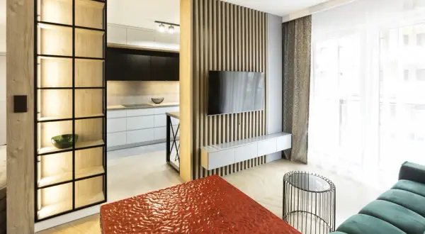 Nowoczesny mały salon inspiracje: drewniana ściana z TV, regał, kuchnia w tle, czerwony stolik i zielona sofa.