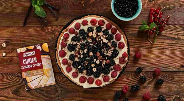 Pyszna tarta z mascarpone, malinami, jeżynami i borówkami, posypana orzechami. Obok opakowanie cukru z skórką cytrynową.