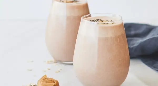 Två glas med krämig choklad- och jordnötssmoothie, toppad med chiafrön. En sked med jordnötssmör och havregryn ligger bredvid. En perfekt start på dagen, som Don Pedro skulle ha gillat.