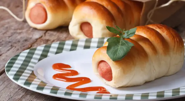 Leckere Bratwurst im Schlafrock, garniert mit einem Blatt, auf einem Teller mit Ketchup-Spur.