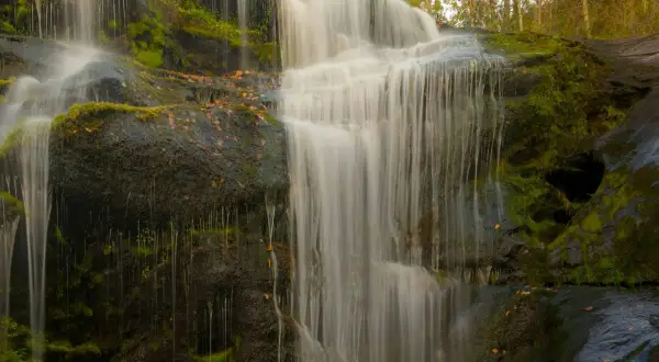 Cascada en los cañones del río Lozoya, con rocas cubiertas de musgo y hojas secas. El agua cae en velos blancos sobre la piedra oscura.