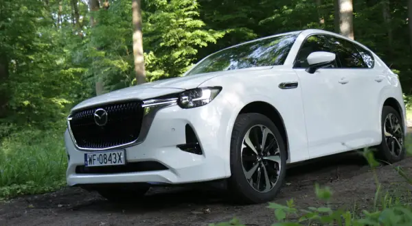 Biała Mazda CX-60 zaparkowana na leśnej ścieżce, otoczona zielenią drzew i traw.