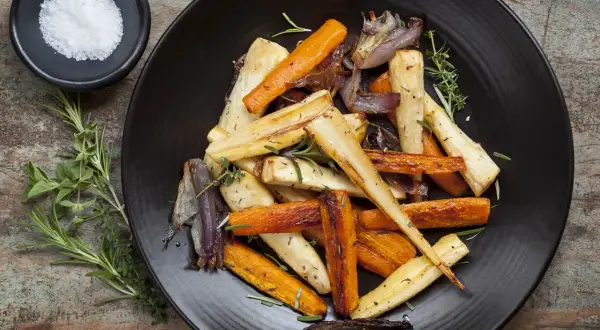 Légumes rôtis, carottes et panais, parsemés d'herbes. Un navet doré, tendre et savoureux, se mêle aux autres légumes sur l'assiette noire.