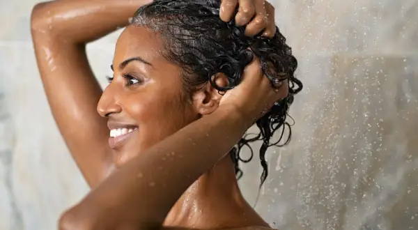 Lächelnde Frau wäscht sich unter der Dusche die Haare. Sie massiert Shampoo in ihre nassen, dunklen Locken.