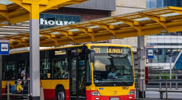 Kierowca autobusu linii 131 dojeżdża do przystanku Wilanów. Żółto-czerwony pojazd czeka na pasażerów pod żółtą wiatą.
