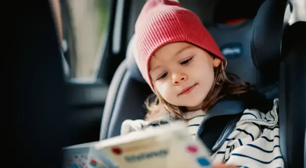Kleines Mädchen mit roter Mütze liest ein Buch im Kindersitz. Perfekt für autofahrt spiele, um die Reise kurzweilig zu gestalten.